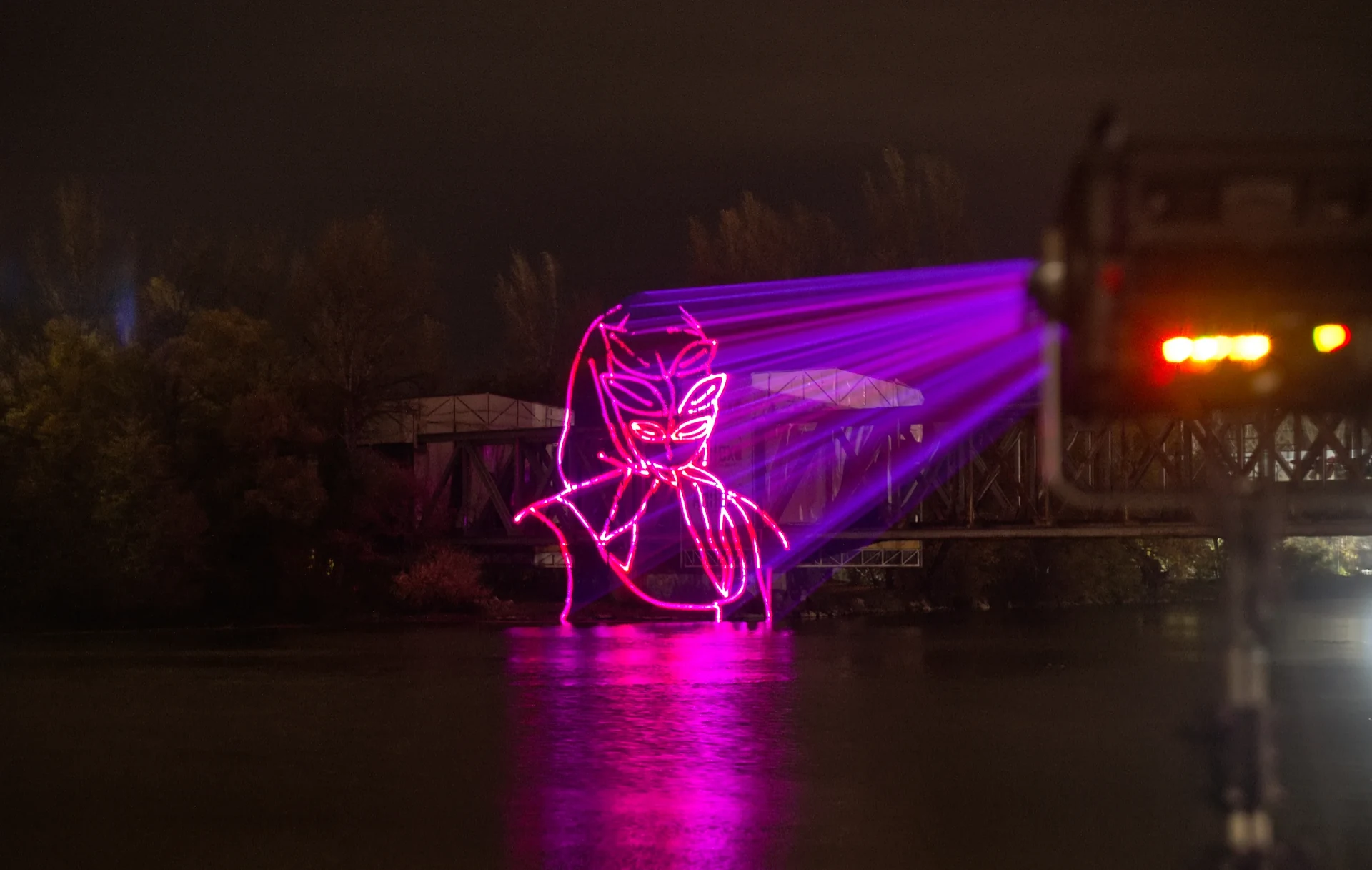 A pink and violet folk figure projected over the river at night